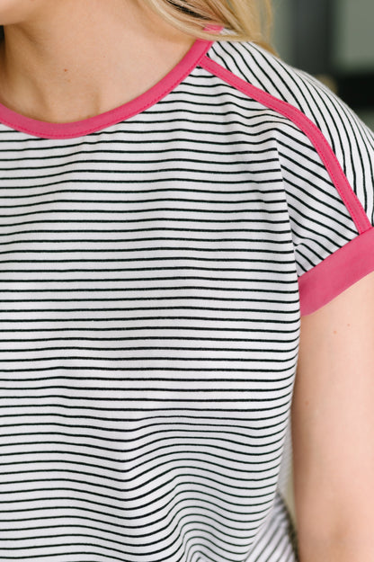 Close-up of a short-sleeve ringer T-shirt with thin black horizontal stripes on a white base and bright pink trim along the crew neck and sleeve cuffs, worn by a model.