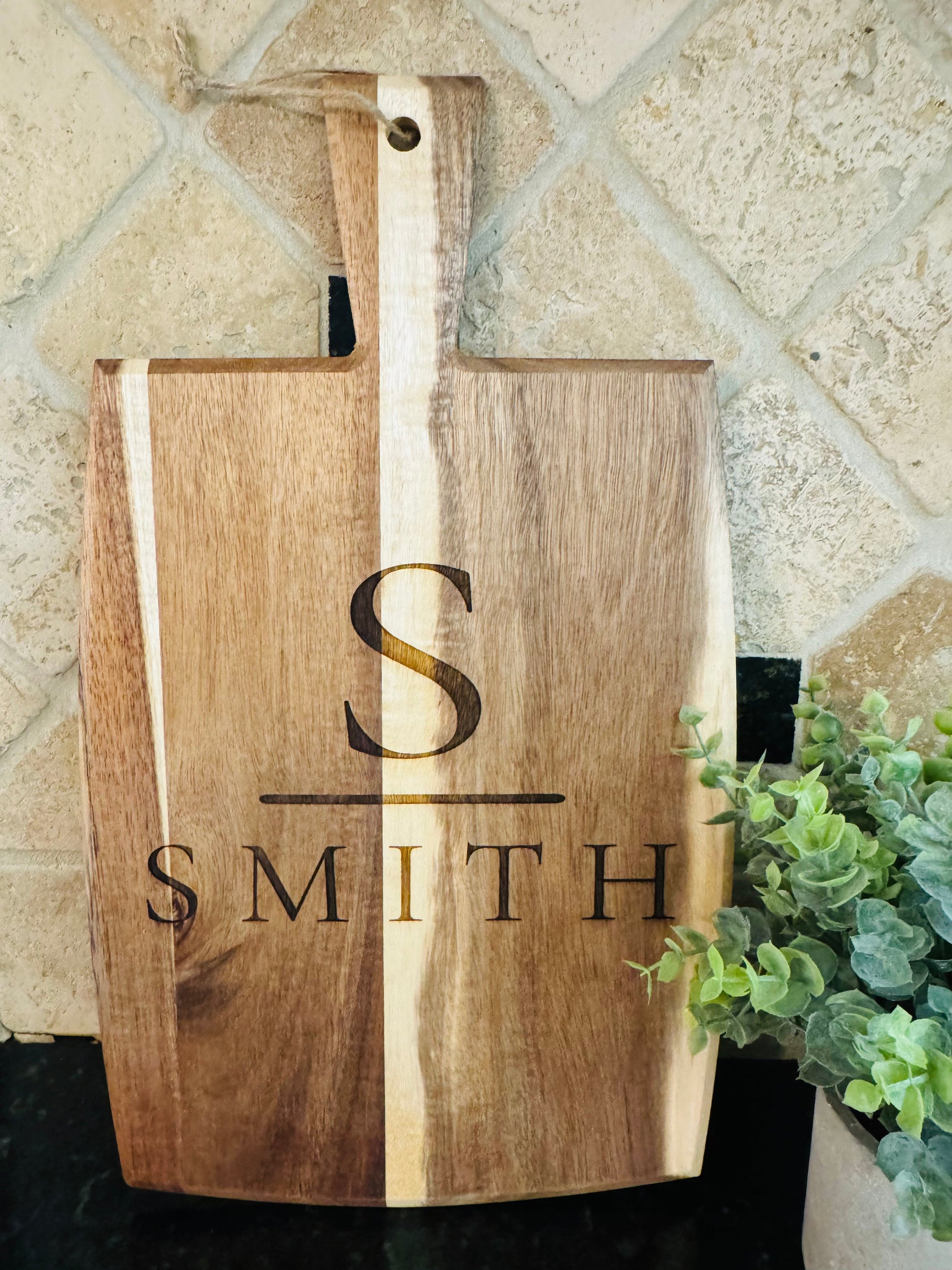 Wooden cutting board with engraved initials and name, placed on a tiled wall.