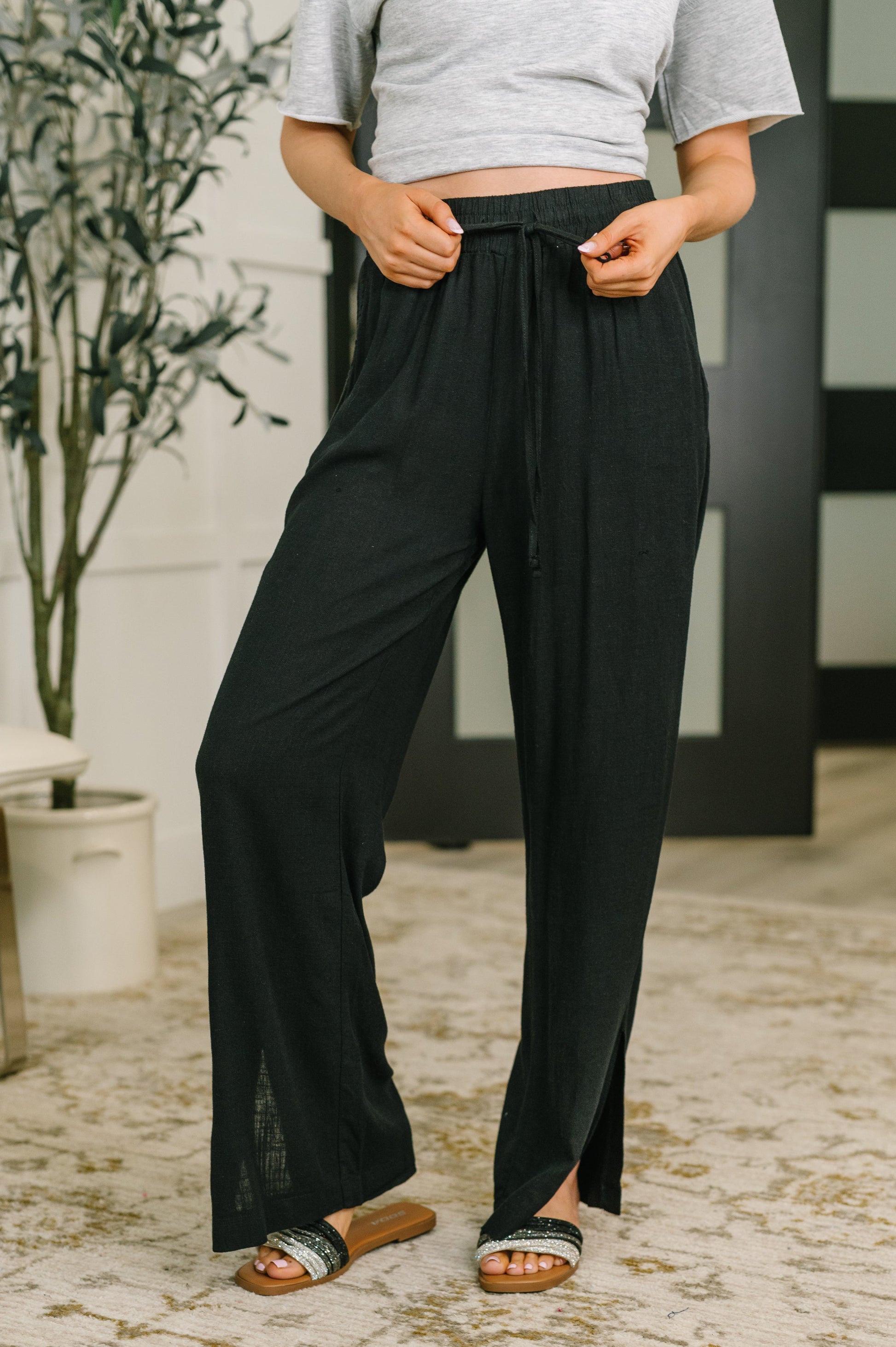 A person wearing loose black wide-leg drawstring pants with side slits, paired with a light gray cropped T-shirt and black slide sandals, standing indoors on a patterned rug.