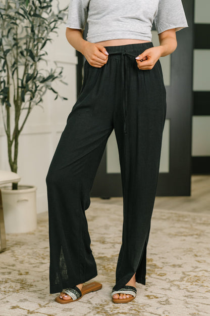 A person wearing loose black wide-leg drawstring pants with side slits, paired with a light gray cropped T-shirt and black slide sandals, standing indoors on a patterned rug.