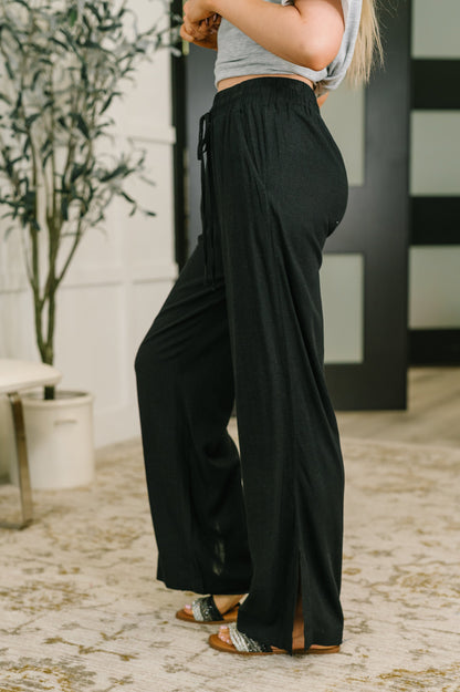 A person wearing loose black wide-leg drawstring pants with side slits, paired with a light gray crop top and patterned sandals, standing indoors on a patterned rug.