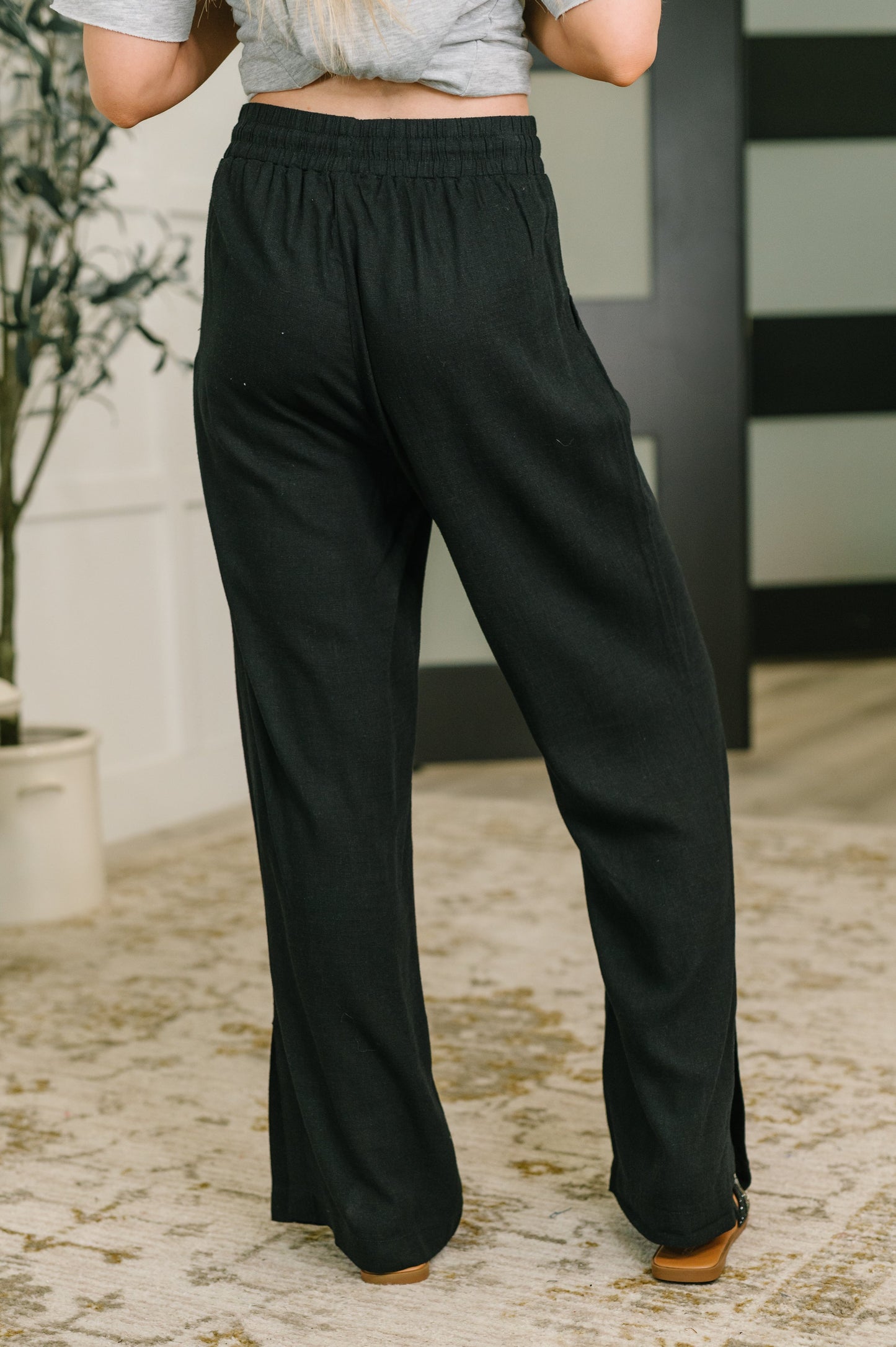 Black wide-leg lounge pants with an elastic waistband, shown on a model from the back in an indoor setting with a patterned rug and neutral wall.