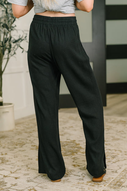 Black wide-leg lounge pants with an elastic waistband, shown on a model from the back in an indoor setting with a patterned rug and neutral wall.