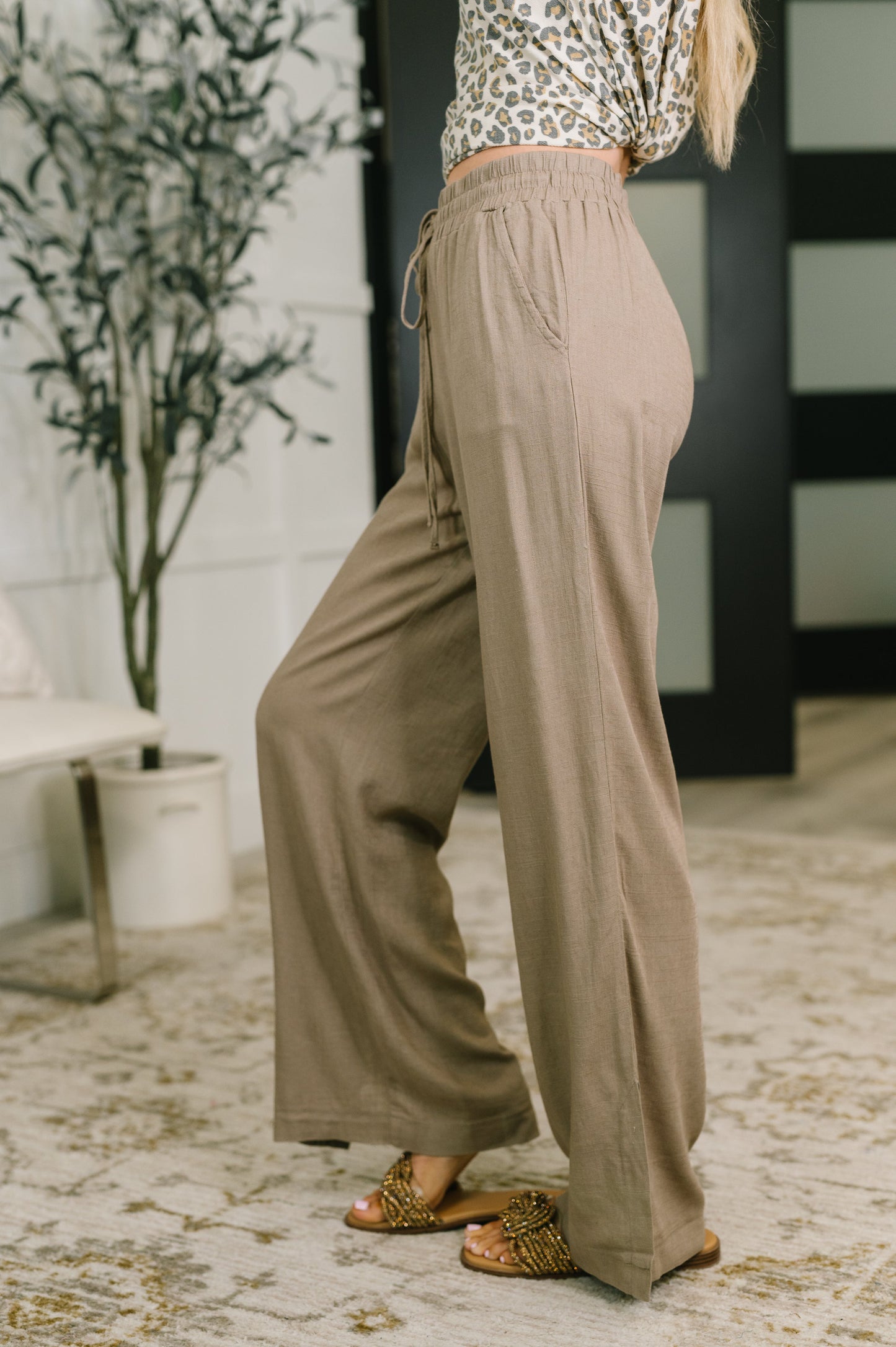 A person wearing loose, wide-leg taupe pants with an elastic drawstring waist, paired with a patterned top and flat sandals, standing indoors on a patterned rug.