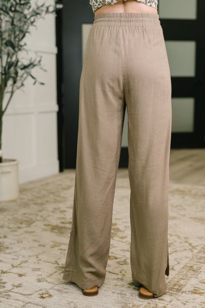 A person shown from the back wearing loose, wide-leg taupe pants in an indoor setting with a patterned rug and dark doorway in the background.