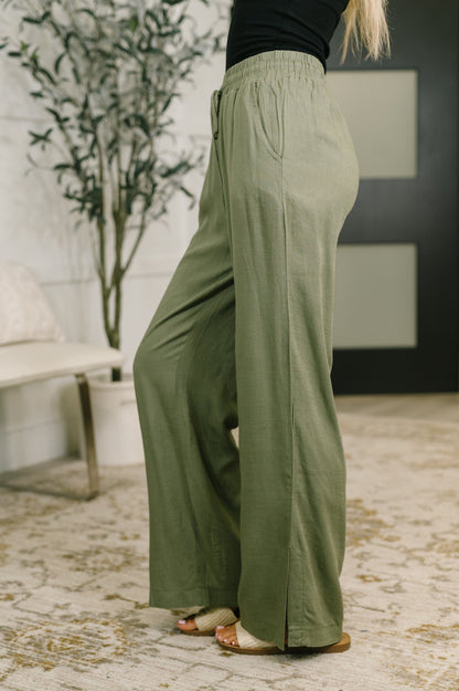 A person wearing wide-leg sage green pants with a black top, standing on a patterned rug in an indoor setting.