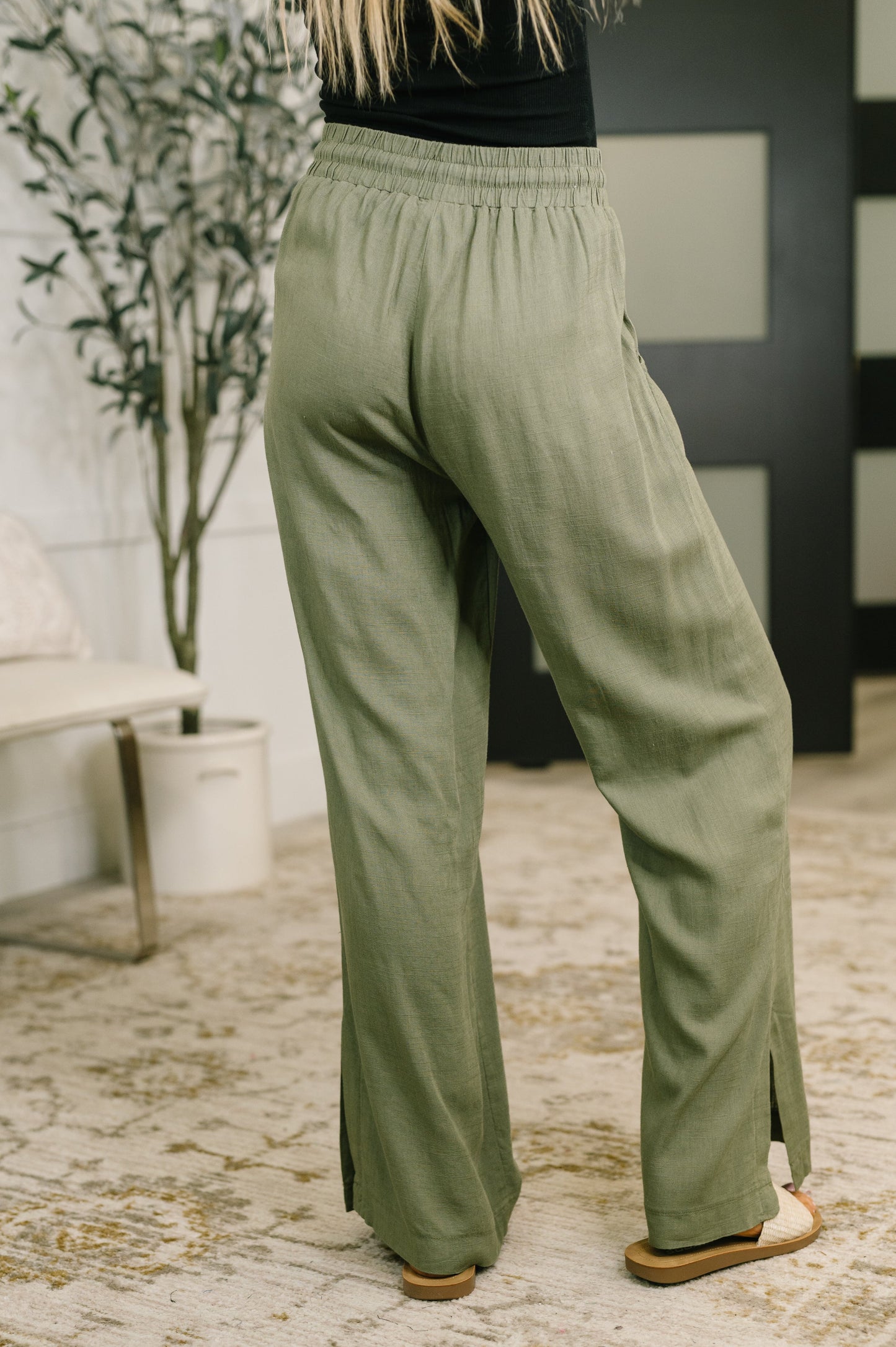 A person wearing loose, wide-leg sage green pants with side slits, shown from the back in an indoor room with a patterned rug, potted plant, and dark paneled wall.