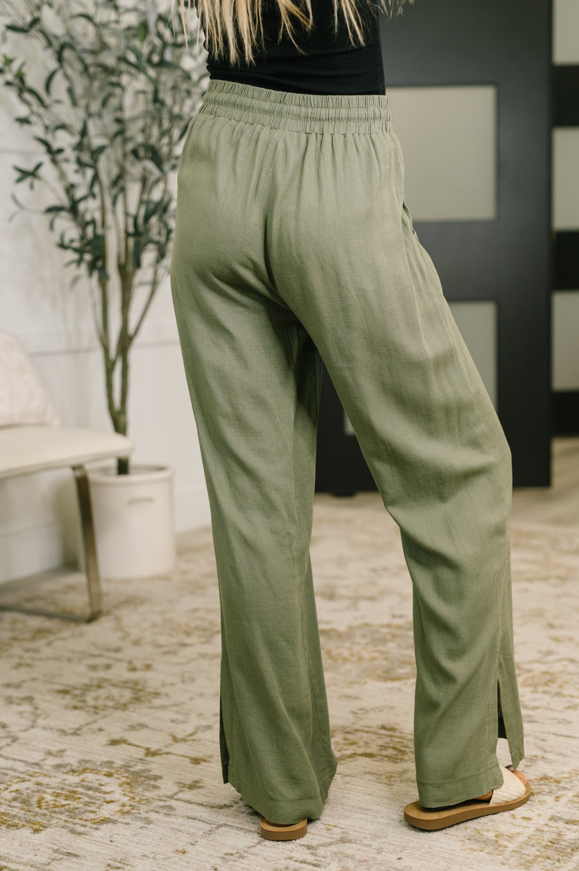 A person wearing loose, wide-leg sage green pants with side slits, shown from the back in an indoor room with a patterned rug, potted plant, and dark paneled wall.