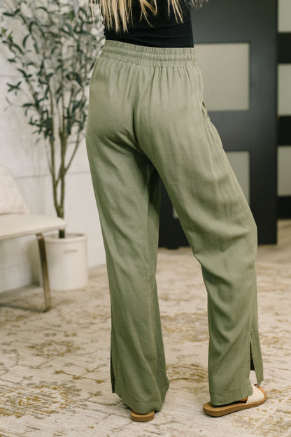 A person wearing loose, wide-leg sage green pants with side slits, shown from the back in an indoor room with a patterned rug, potted plant, and dark paneled wall.