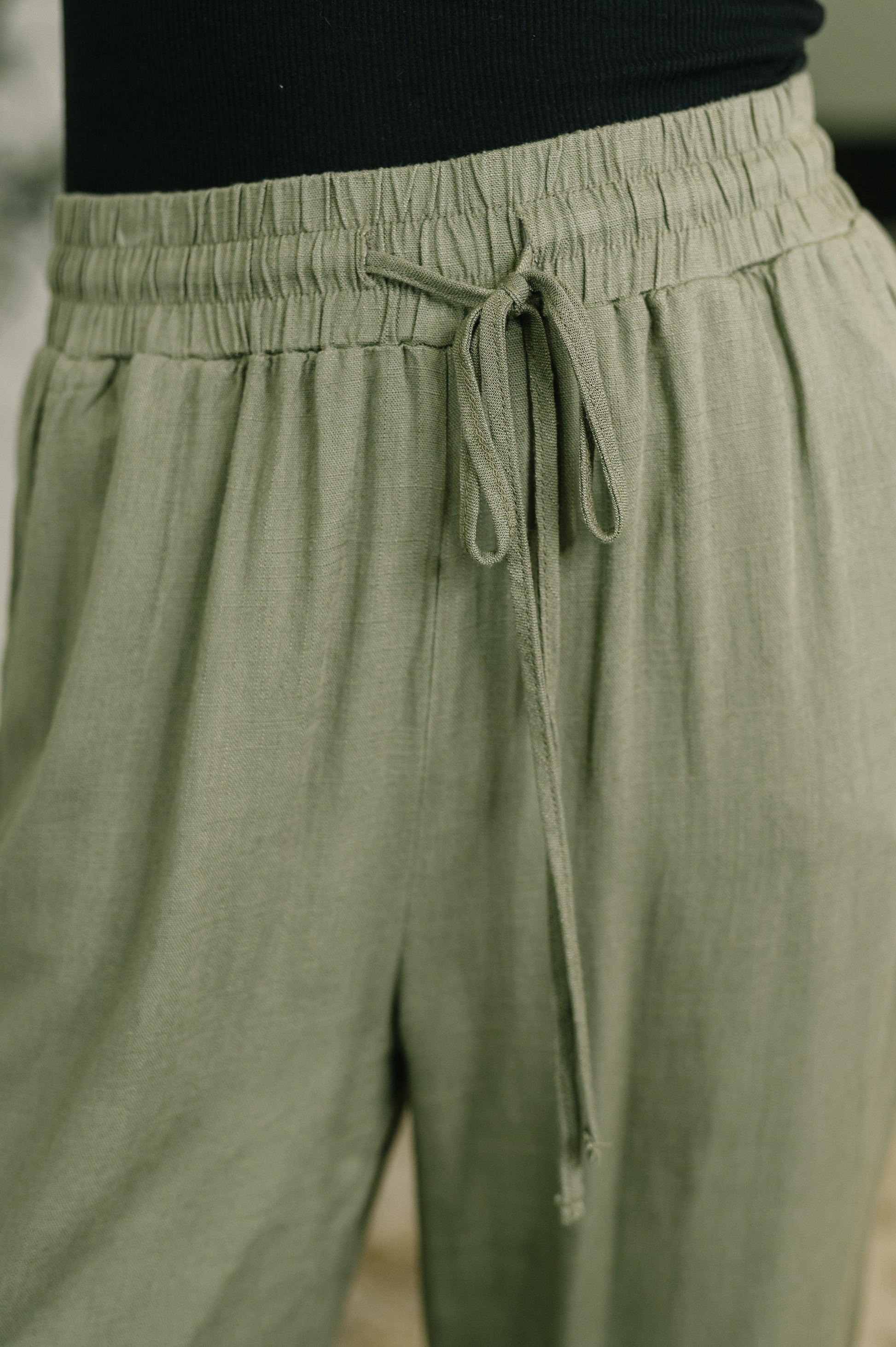Close-up of a person wearing loose sage green drawstring pants with an elastic waistband and black top.