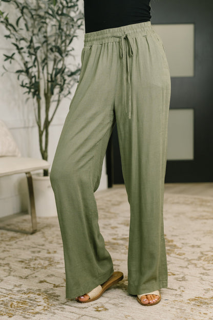 A person wearing loose, wide-leg sage green drawstring pants with a black top and tan slide sandals, standing indoors on a patterned rug near a plant.