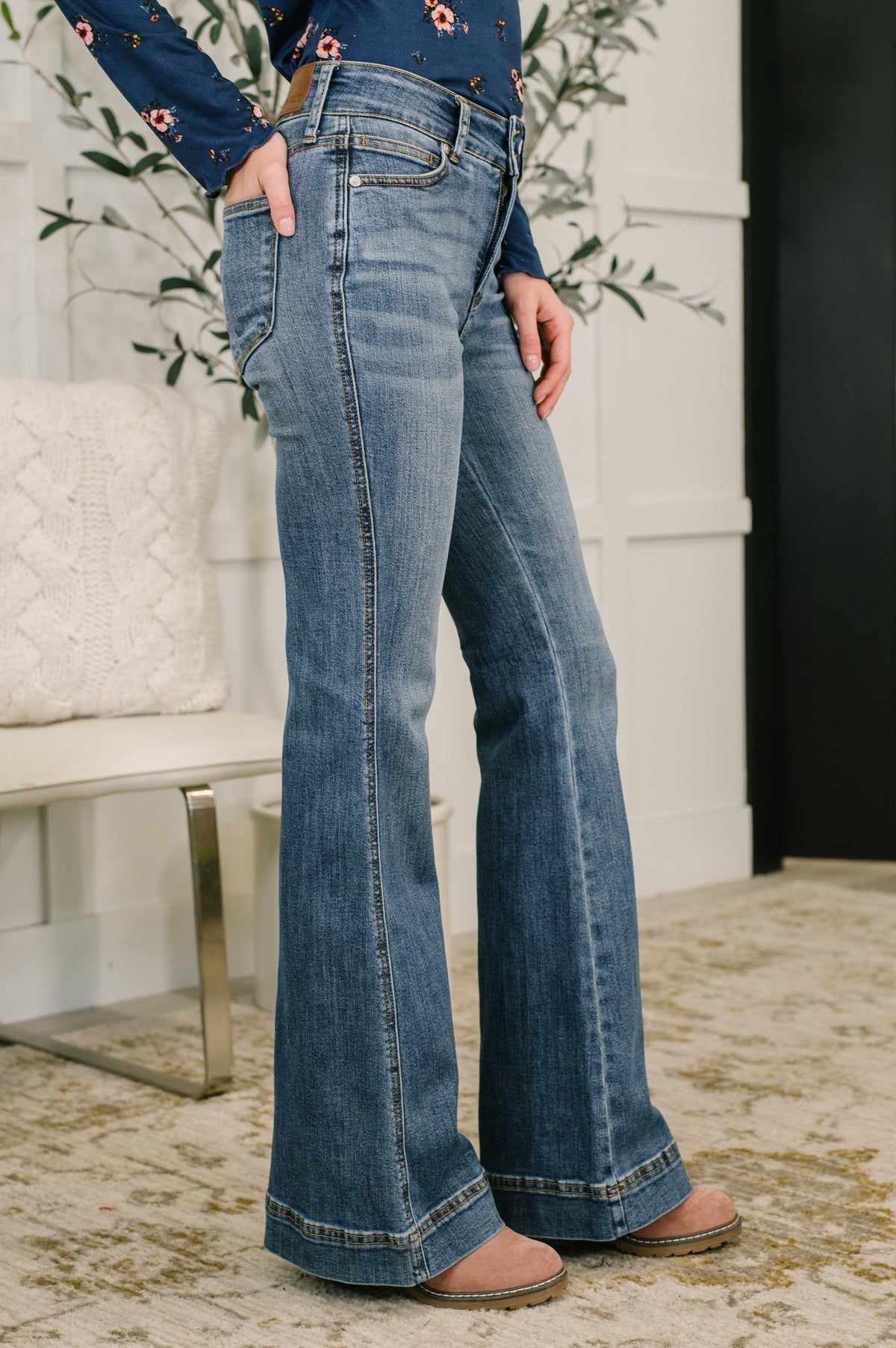 Person wearing blue flared jeans standing indoors with a white wall and plant in the background.