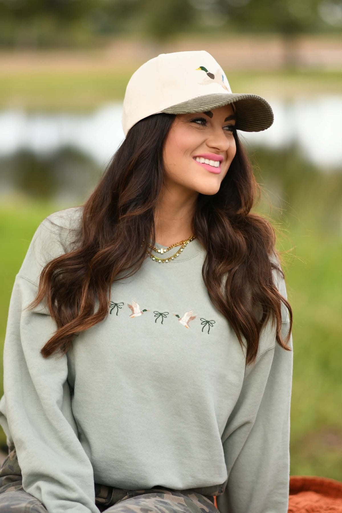 A woman wearing a sage green crewneck sweatshirt with embroidered birds and a beige baseball cap, photographed outdoors in front of a blurred natural background.