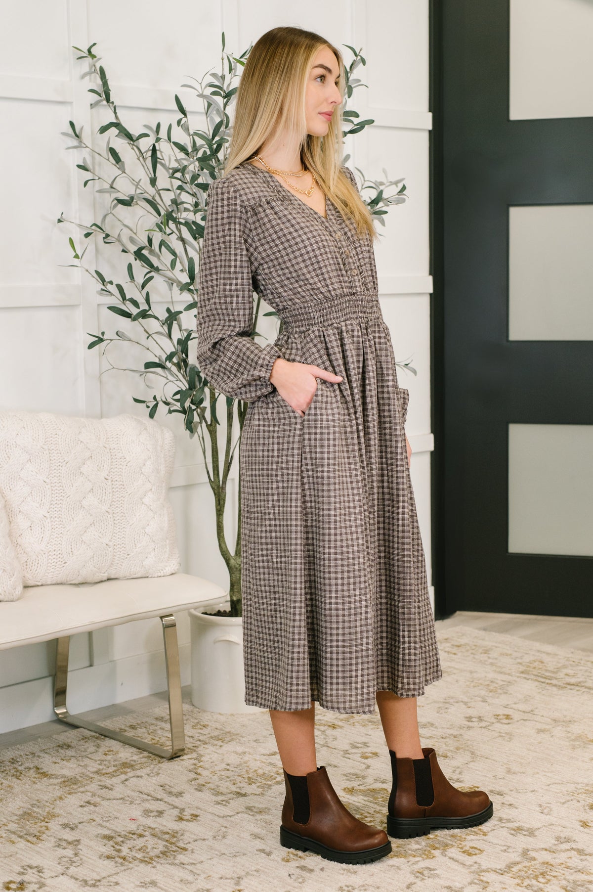 Woman wearing a plaid dress standing in a room with a plant and white wall.