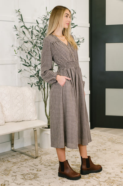 Woman wearing a plaid dress standing in a room with a plant and white wall.