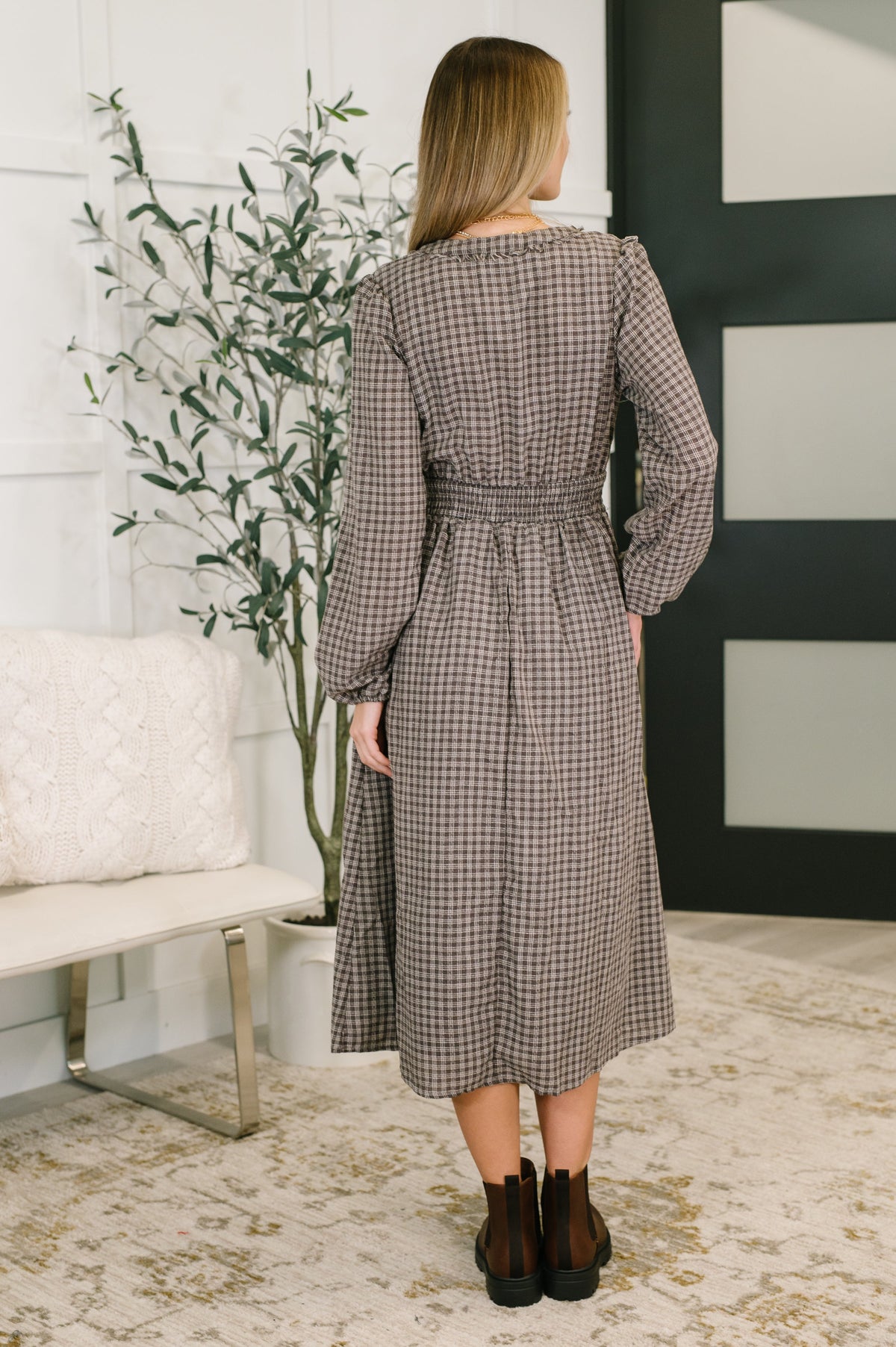 Woman wearing a checkered dress standing in a room with a plant and a bench.