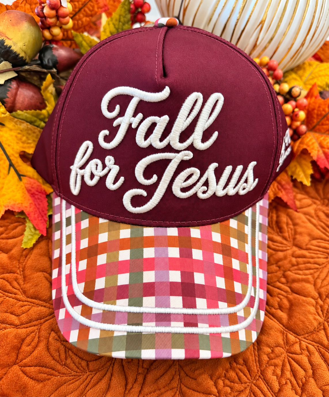 A baseball cap with a maroon front panel and white embroidered text reading 'Fall for Jesus,' paired with a multicolor plaid brim. The cap is styled with autumn leaves, acorns, and pumpkins on an orange quilted blanket.