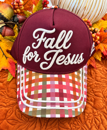 A baseball cap with a maroon front panel and white embroidered text reading 'Fall for Jesus,' paired with a multicolor plaid brim. The cap is styled with autumn leaves, acorns, and pumpkins on an orange quilted blanket.