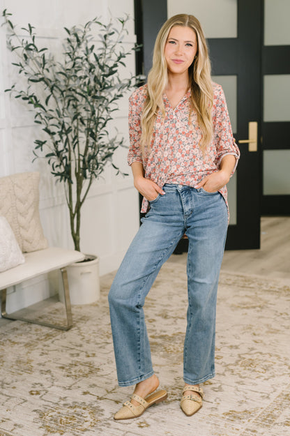 A woman modeling a pink and white floral print blouse with blue straight-leg jeans and tan pointed flats, standing in a bright, modern room with a bench, potted plant, and patterned rug.