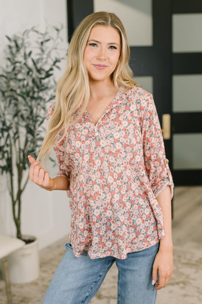 A woman with long blonde hair wearing a loose, three-quarter sleeve floral blouse and blue jeans, standing in a brightly lit indoor setting with a potted plant and a modern black door in the background.
