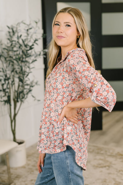 A woman modeling a loose, long-sleeve button-up blouse with a dense small floral print in coral, pink, and blue on a light background, paired with blue jeans in an indoor setting.