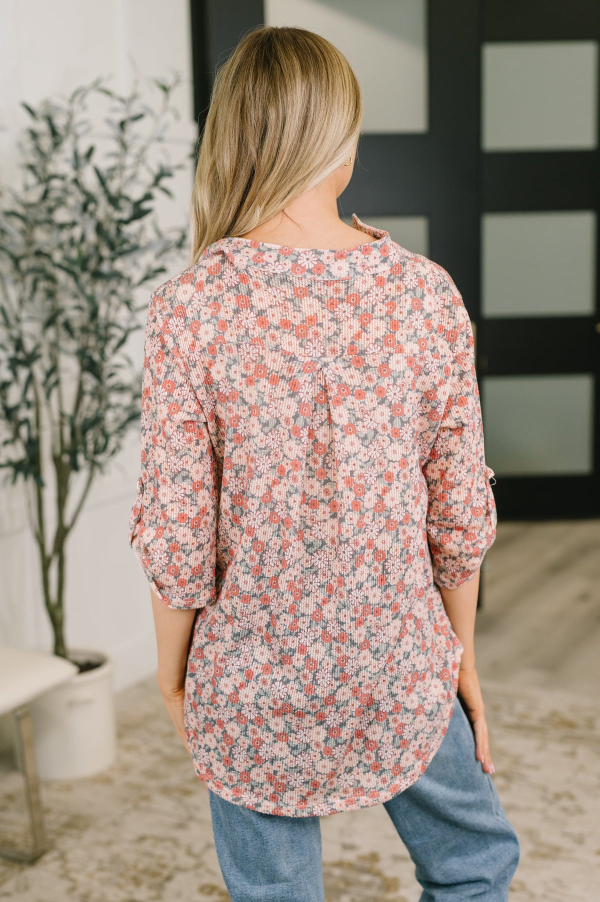 A woman viewed from behind wearing a loose floral long-sleeve top with blue jeans, standing in a modern indoor setting with a plant and dark door in the background.