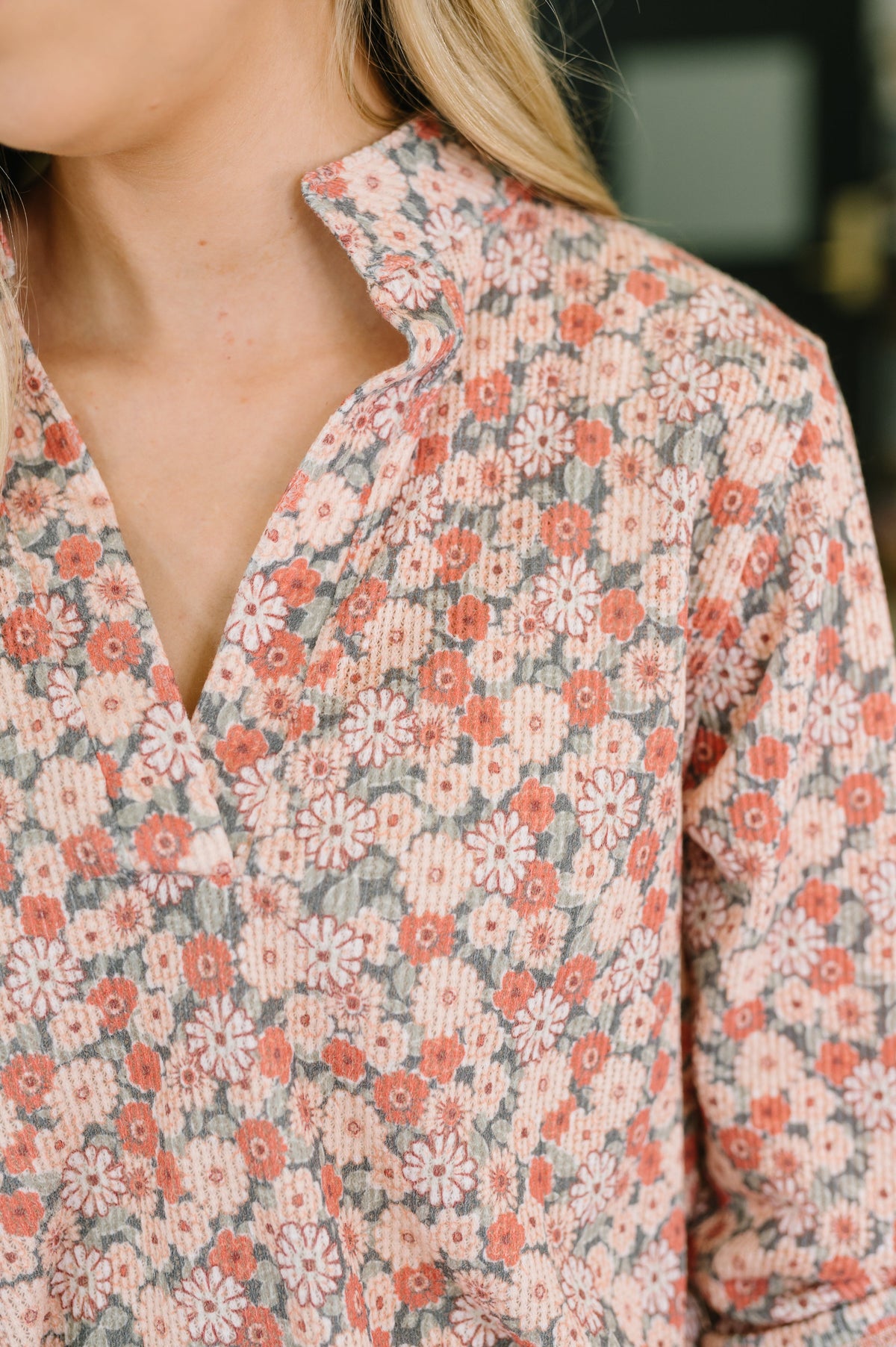 Close-up of a person wearing a long-sleeve collared shirt with a dense floral pattern in coral pink, white, and muted gray-green on a light beige base.