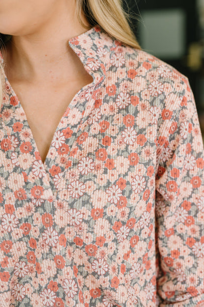 Close-up of a person wearing a long-sleeve collared shirt with a dense floral pattern in coral pink, white, and muted gray-green on a light beige base.