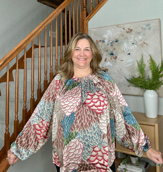 Woman wearing a colorful floral blouse standing in a home setting with a staircase and artwork in the background.
