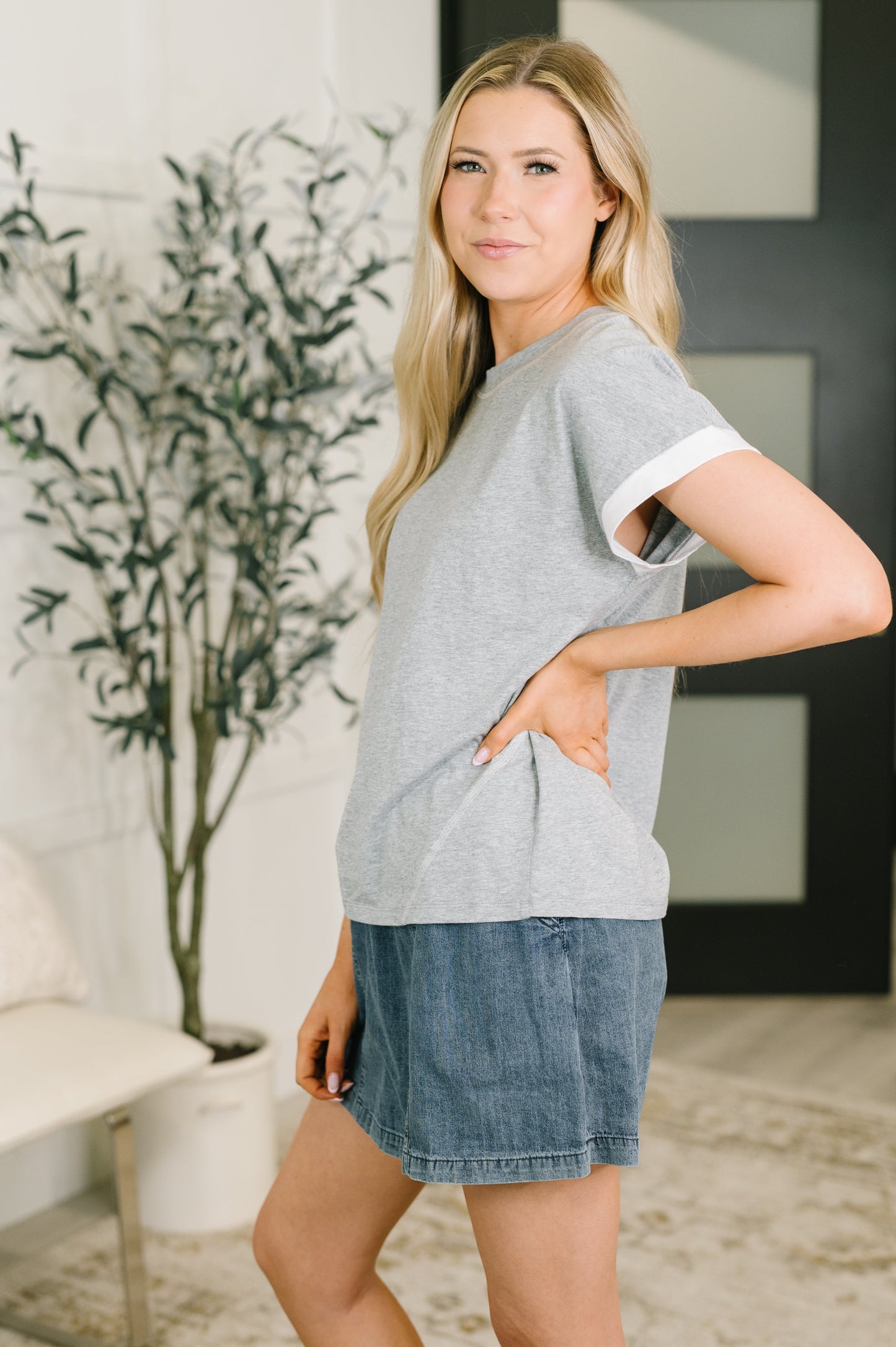 A woman modeling a light gray heather ringer t-shirt with white trim on the sleeves, paired with denim shorts in an indoor setting.