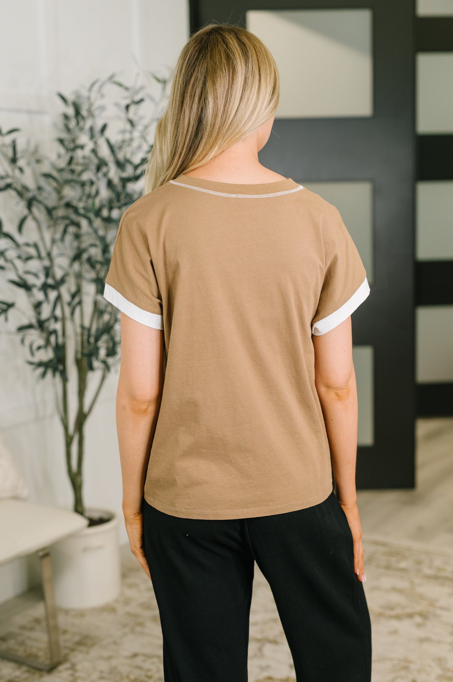 A woman viewed from behind wearing a tan short-sleeve t-shirt with white contrast trim on the collar and sleeve cuffs, paired with black pants in an indoor setting.