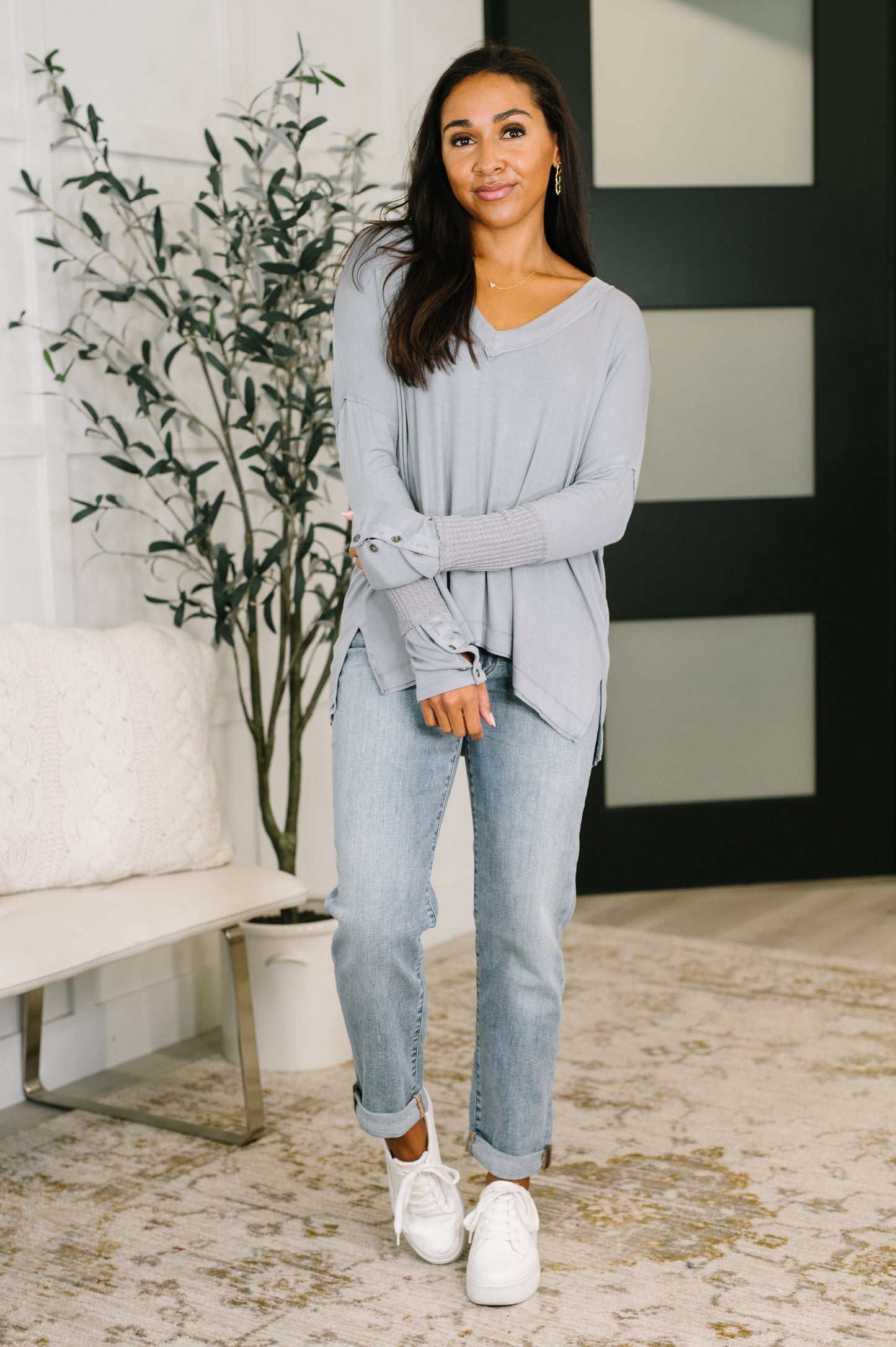 Woman wearing a light gray sweater and jeans standing in a room with a plant and sofa.