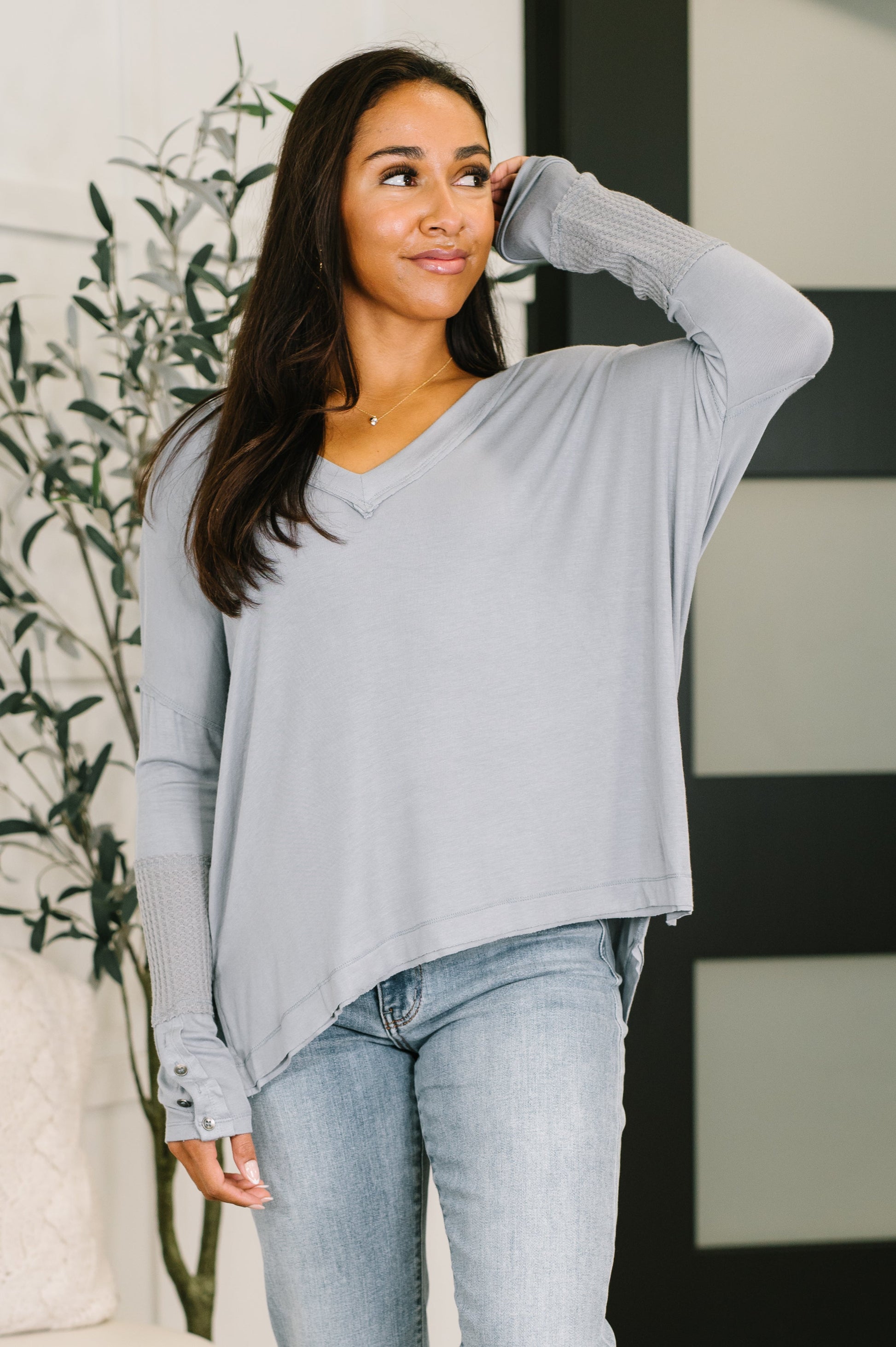 A woman wearing a light gray long-sleeve V-neck top with ribbed cuffs and a relaxed fit, paired with light-wash jeans, standing indoors near a plant and a modern paneled wall.