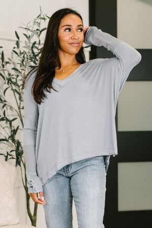 A woman wearing a light gray long-sleeve V-neck top with ribbed cuffs and a relaxed fit, paired with light-wash jeans, standing indoors near a plant and a modern paneled wall.