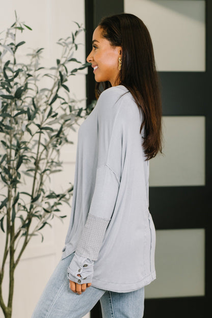 Woman wearing a light gray sweater and jeans standing next to a plant indoors.