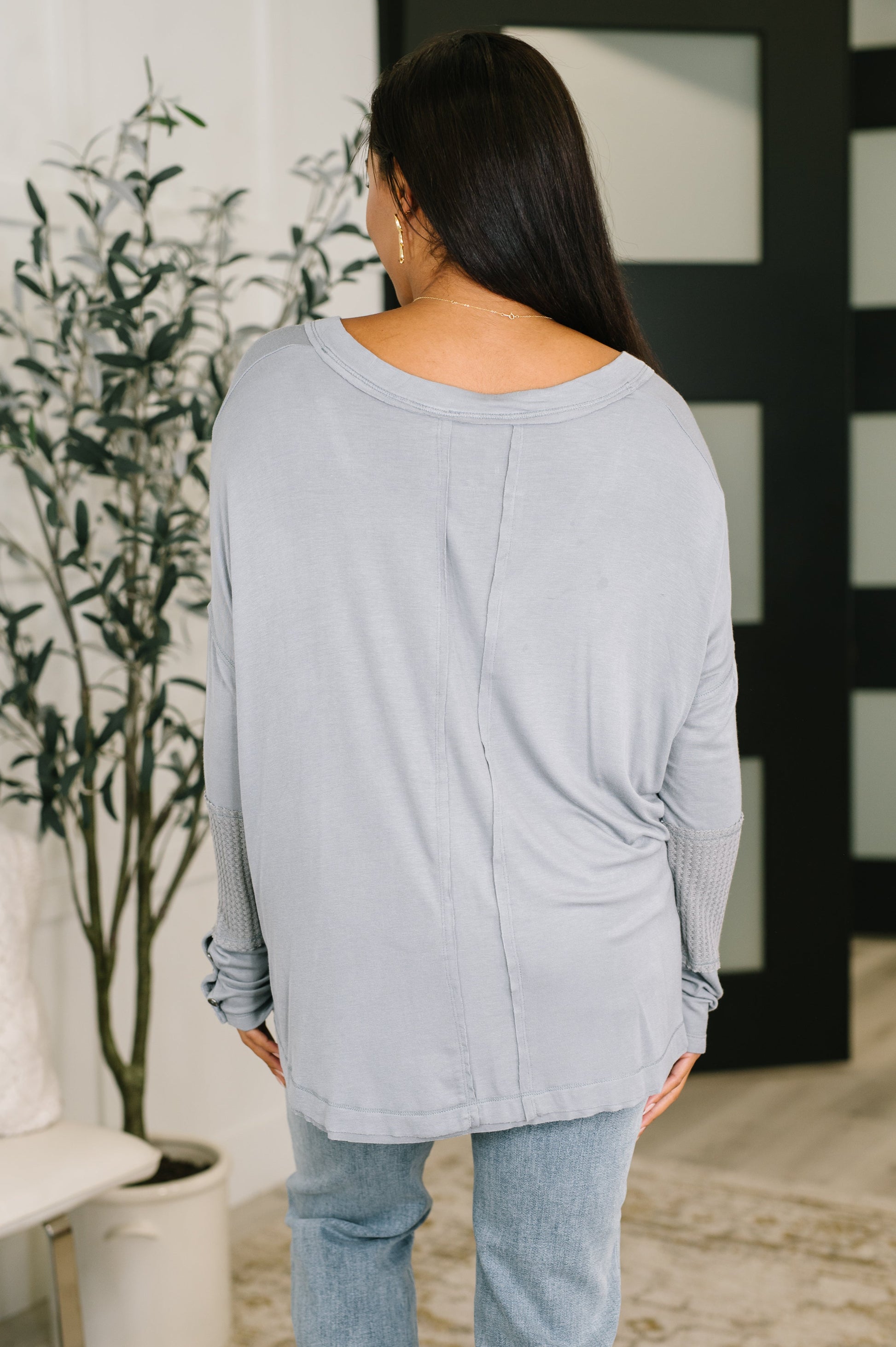 woman in grey scoop neck long sleeve top facing backwards to show longer length of top. 