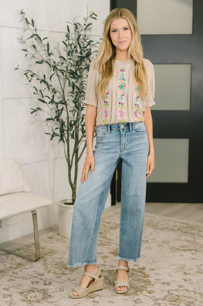 Woman wearing an embroidered top and jeans in a room with a plant and furniture.