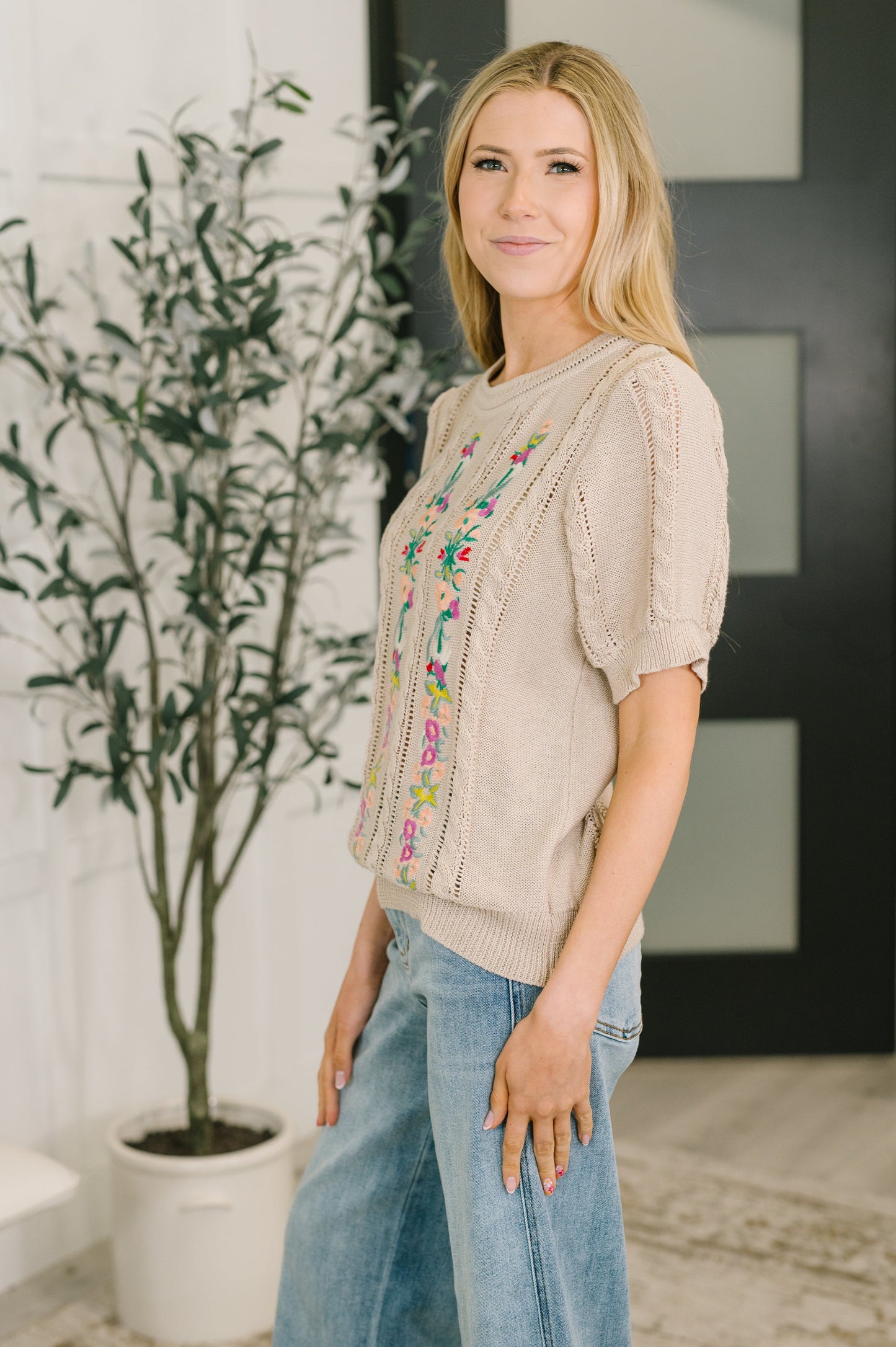Woman wearing a beige sweater with colorful embroidery and blue jeans standing indoors.