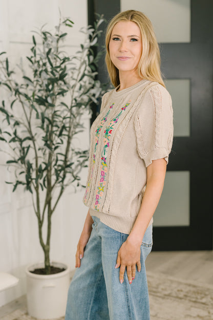 Woman wearing a beige sweater with colorful embroidery and blue jeans standing indoors.