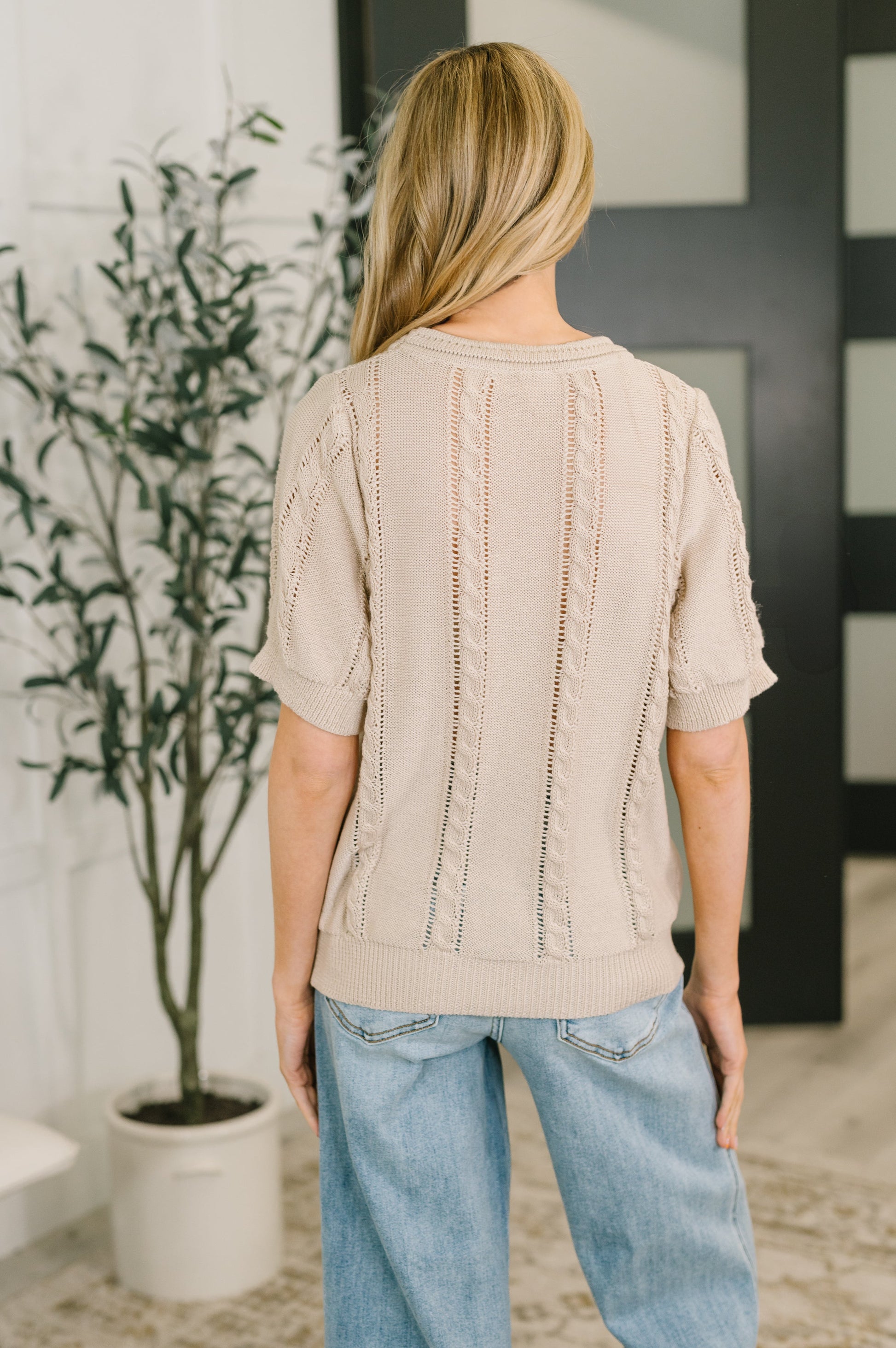 Person wearing a beige textured sweater and blue jeans standing in a room with a plant and door in the background.