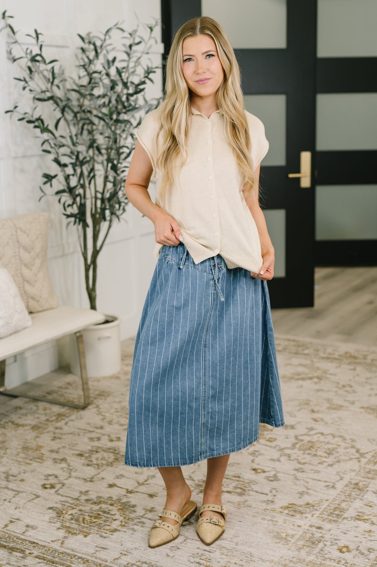 A woman modeling a cream short-sleeve button-up shirt and a blue pinstriped midi skirt, paired with gold flat shoes, standing in a modern living room setting.