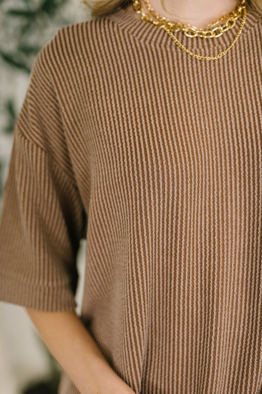 Person wearing a brown ribbed sweater with gold chain necklaces.
