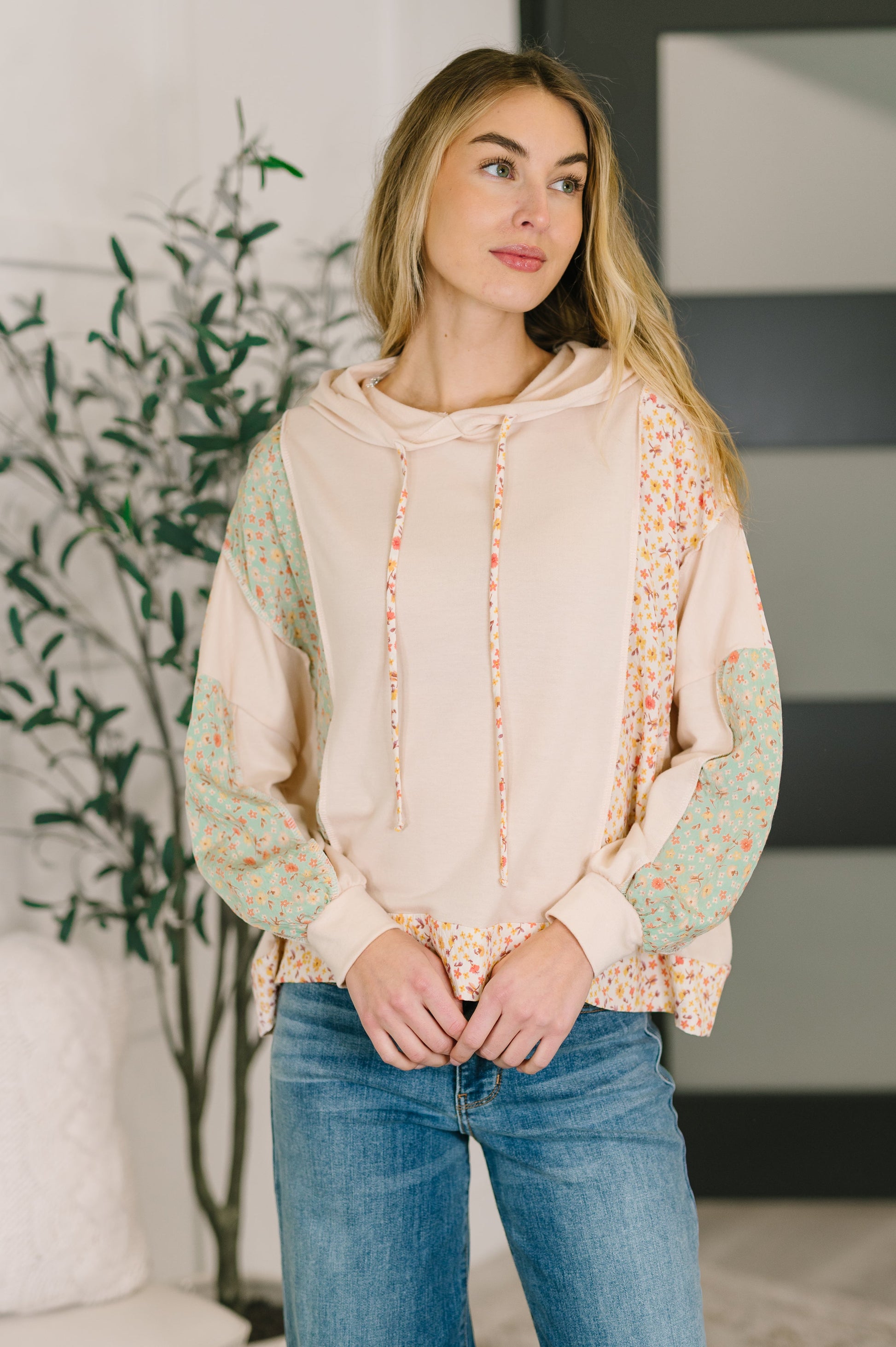 Woman wearing a beige hoodie with colorful sleeves in a room with a plant and a couch.