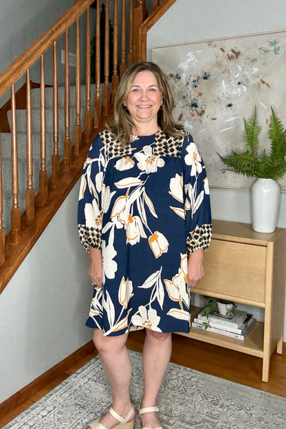 Woman in a floral dress standing in a home setting with a staircase and bookshelf.