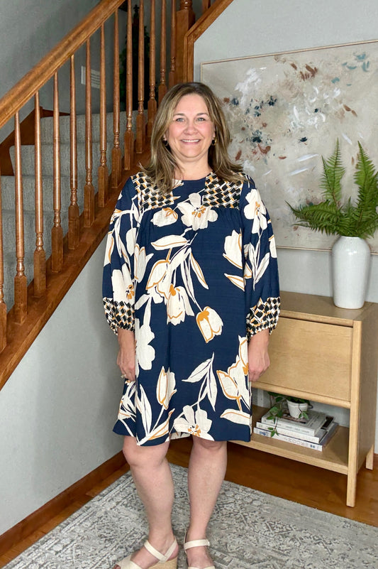 Woman in a floral dress standing in a home setting with a staircase and bookshelf.
