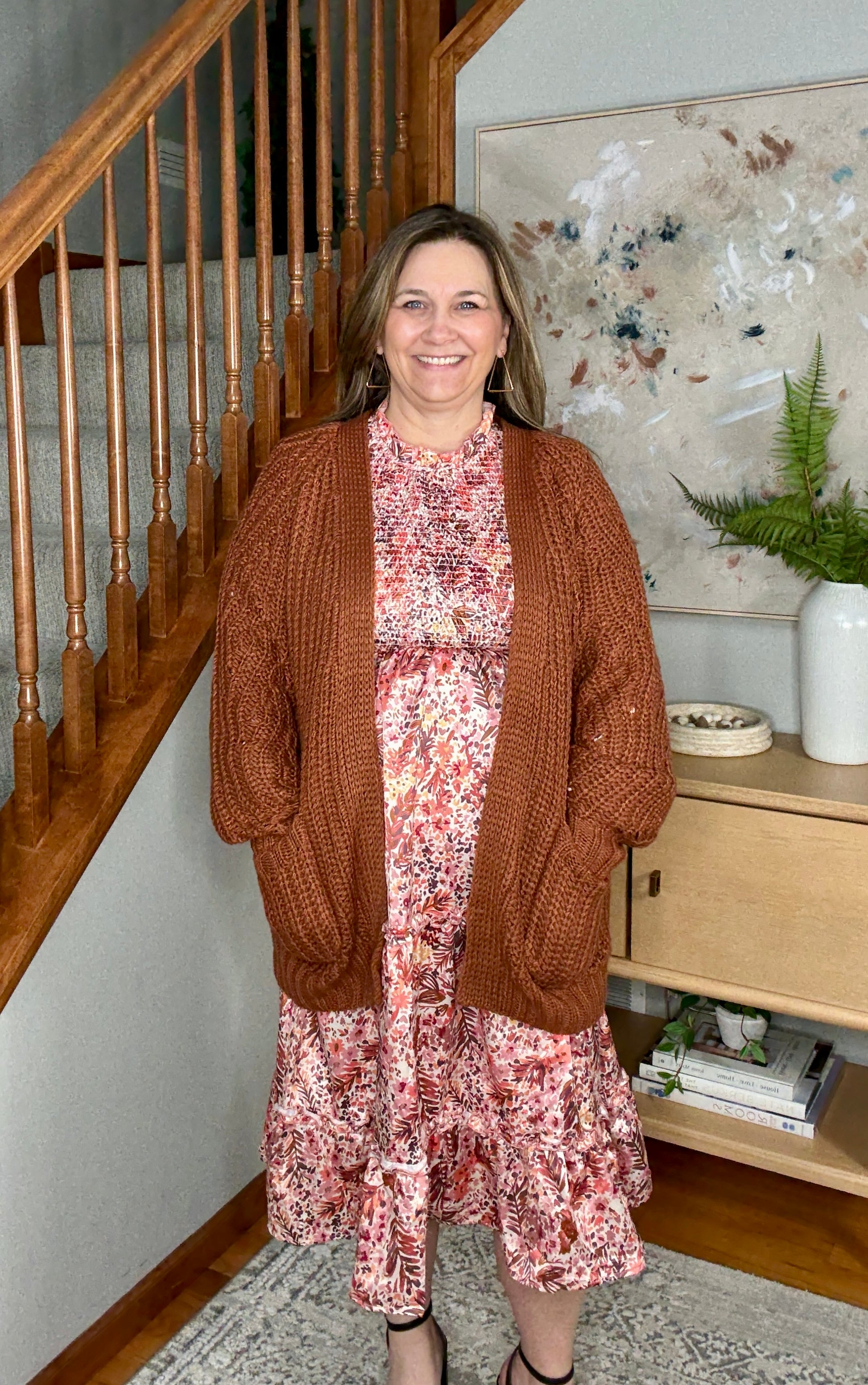 Woman wearing a brown cardigan over a floral dress in a home setting.