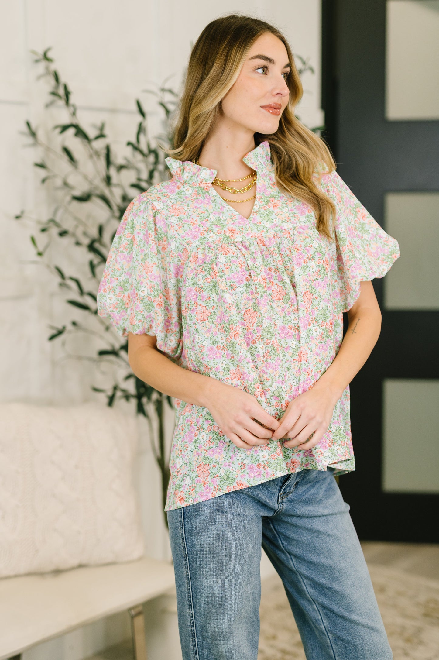 Woman wearing a floral blouse and jeans standing in a room with a plant and furniture.