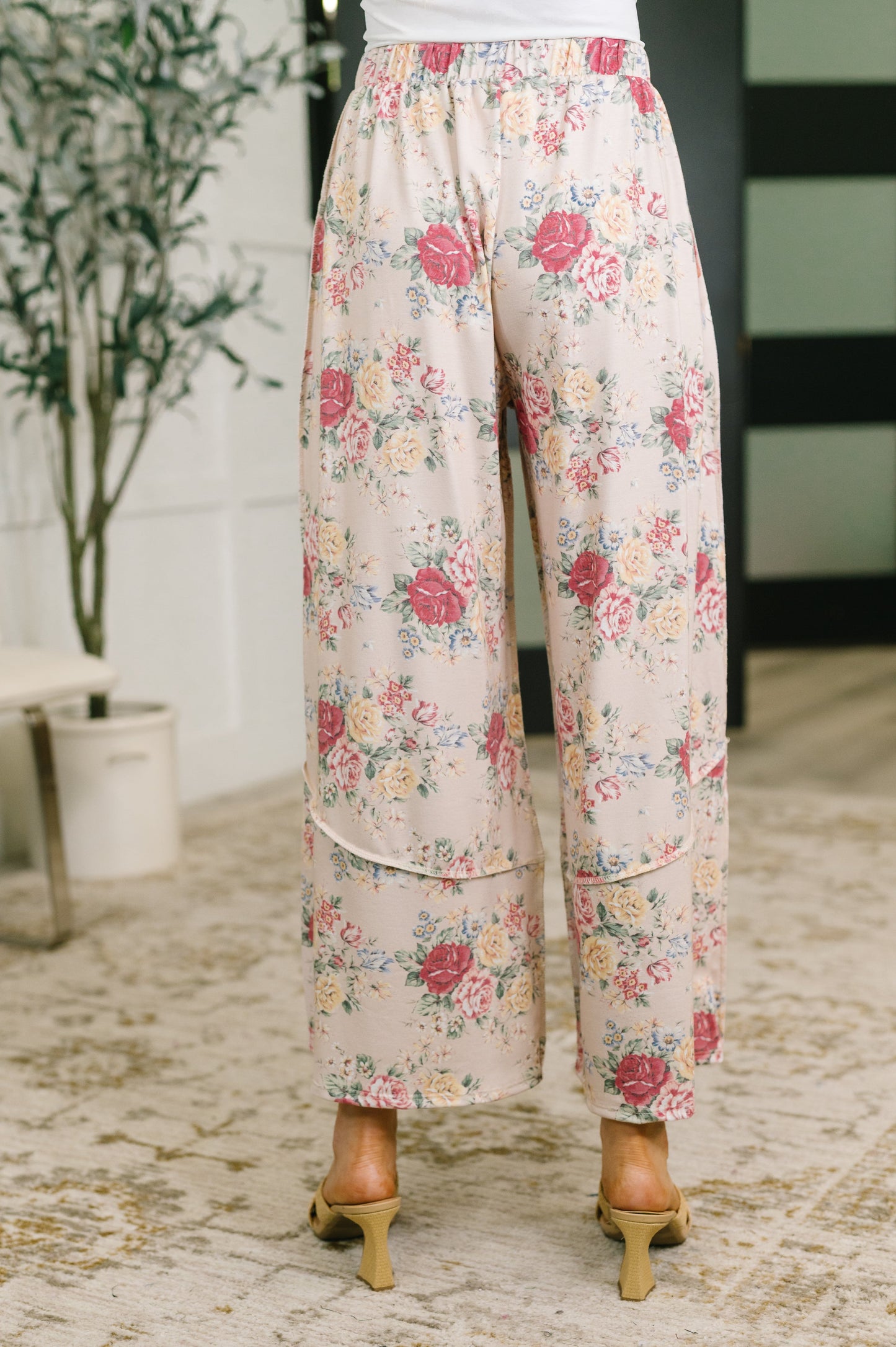 A person shown from the waist down wearing wide-leg cropped pants with a pink, red, and yellow floral print on a light beige background, paired with tan heeled sandals in an indoor setting.