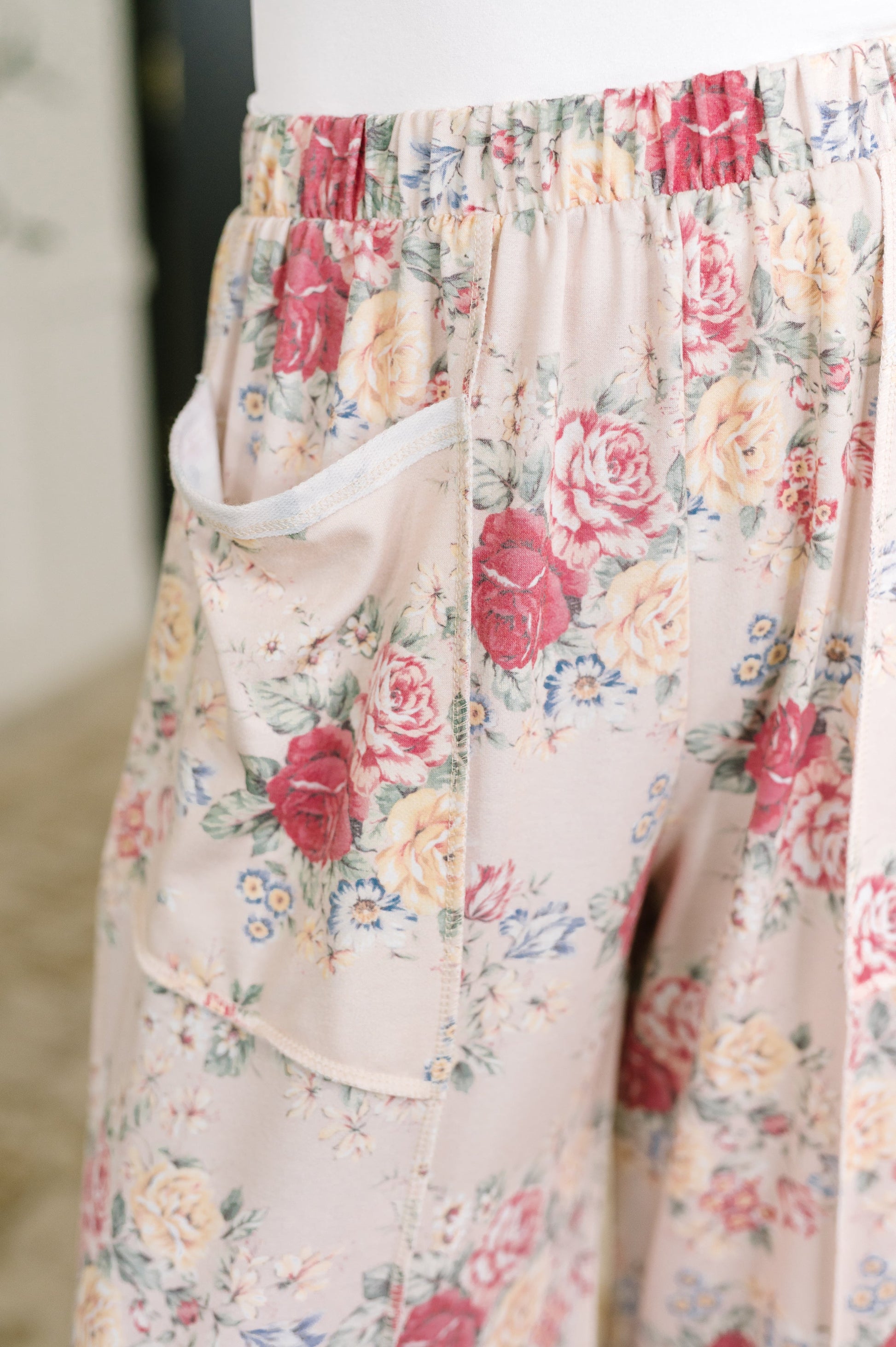 Close-up of a pair of floral print pants with an elastic waistband and a side pocket, featuring a pink, red, and cream rose pattern on a light beige background.