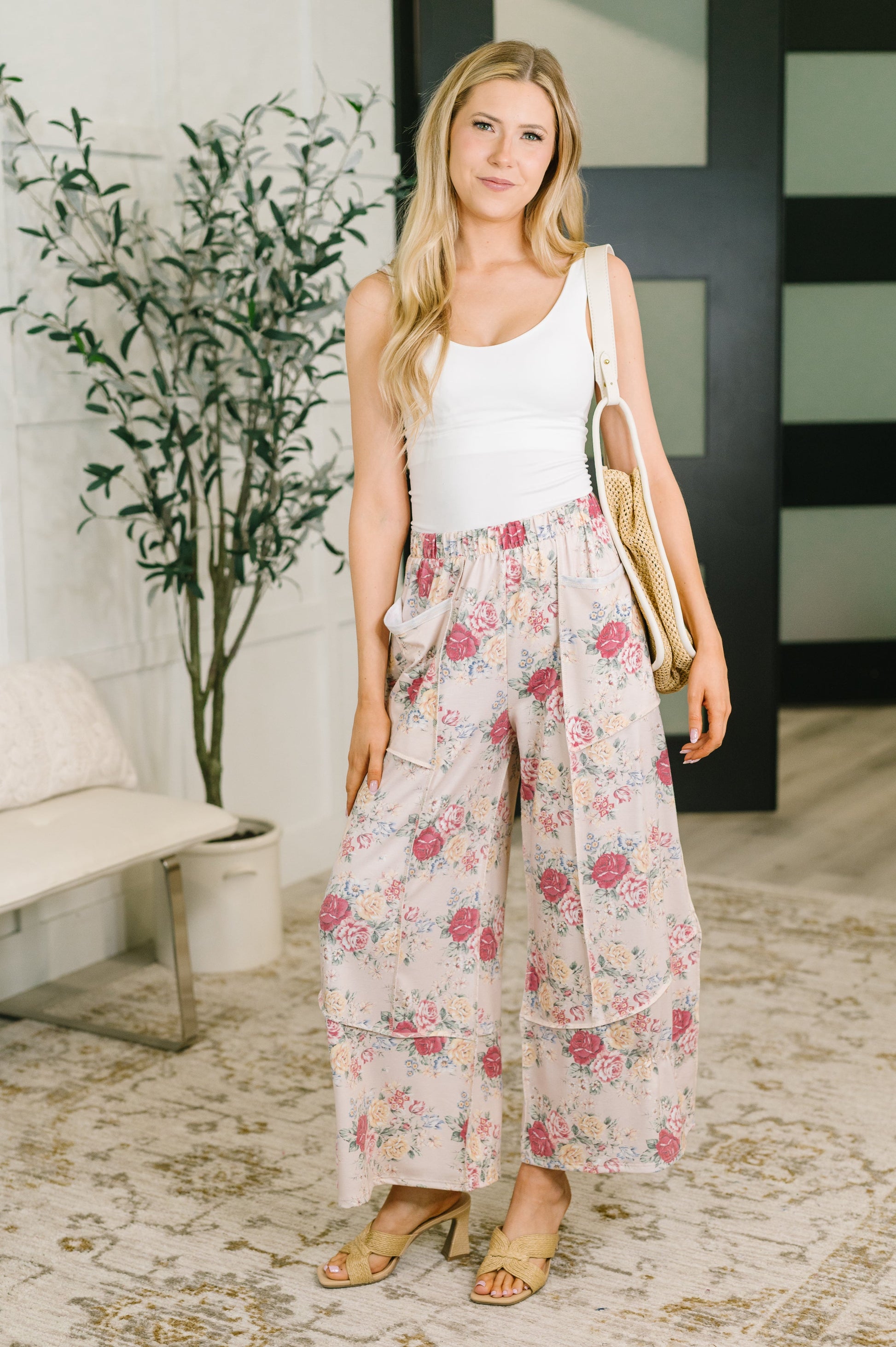 A woman stands indoors wearing a white sleeveless top and loose, wide-leg floral pants with pink and red flowers on a light background. She is holding a tan shoulder bag and wearing beige heeled sandals.